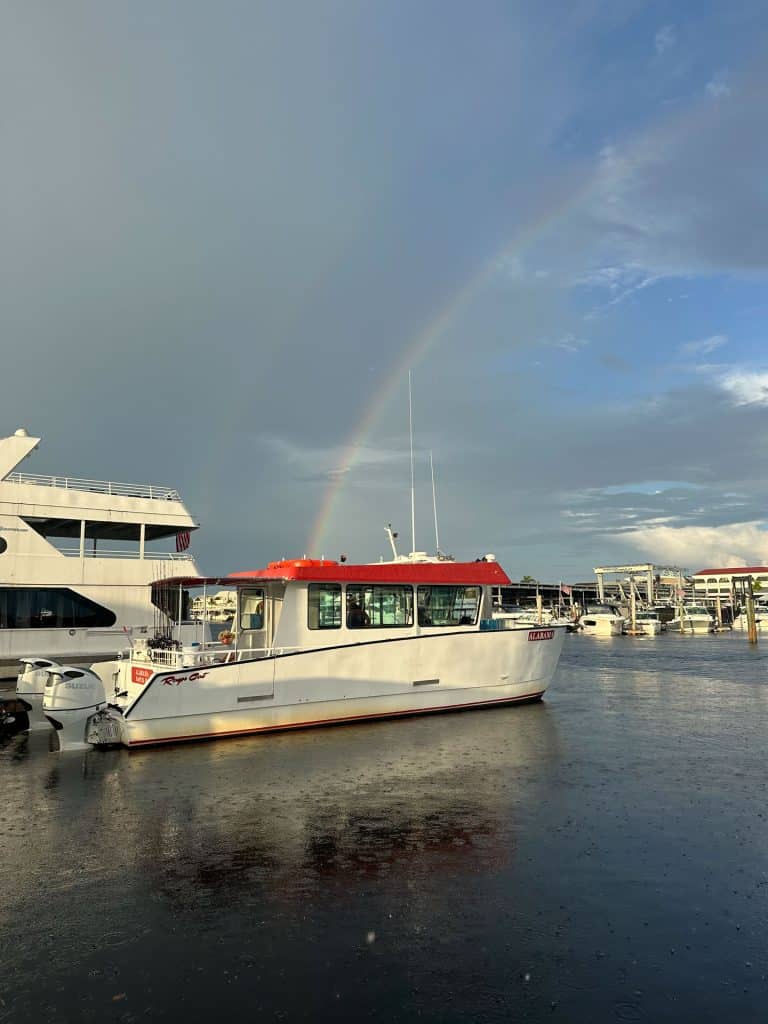 A white boat with a red roof is docked on calm water, with a faint rainbow arching across a partly cloudy sky. Other boats and a marina are visible in the background, creating the perfect setting for an unforgettable Naples Fishing Charter adventure.