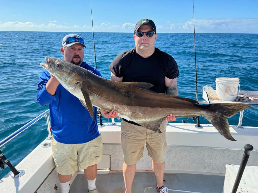Two men on a boat holding a large, freshly caught fish, with the blue ocean and sky in the background—a memorable moment from an exciting Naples FL Deep Sea Fishing adventure. One man wears a blue shirt and sunglasses, while the other sports a black shirt and cap, making this Naples Fishing Charter trip truly unforgettable.