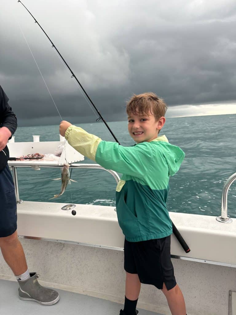 Smiling child in a green rain jacket holds up a small fish on a fishing pole while standing on a boat during an exciting Naples FL Deep Sea Fishing adventure. The ocean and dark, cloudy sky are visible in the background.