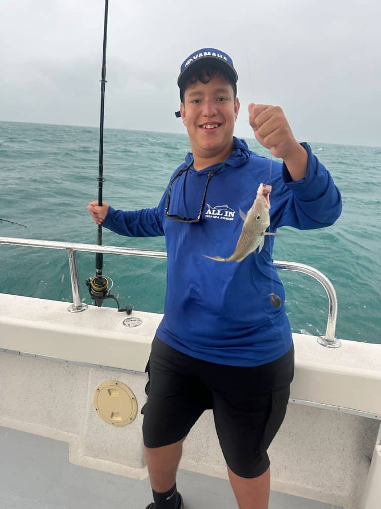 Smiling boy wearing a blue shirt and cap stands on a boat during a Naples Fishing Charter, holding a fishing rod and proudly displays a small fish he caught, with the ocean in the background.