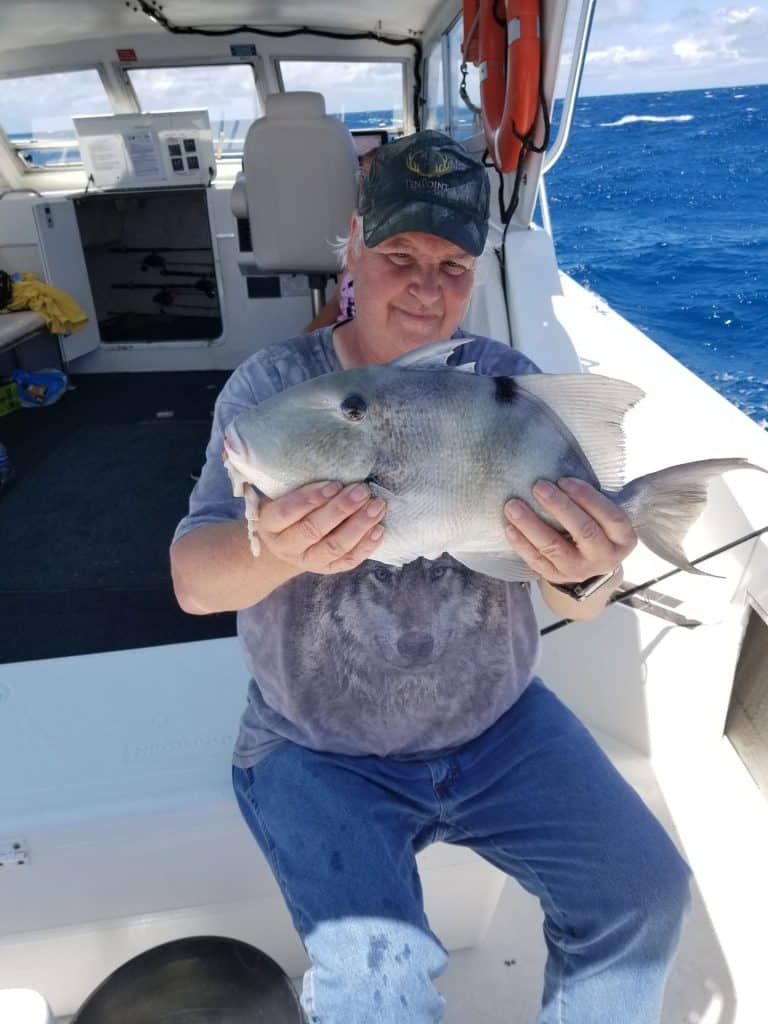 Person sitting on a boat holding a large gray fish with both hands during an exciting Naples FL Deep Sea Fishing adventure. The ocean is visible in the background, and some boat equipment can be seen inside the cabin. The person, dressed in a cap and jeans, showcases the kind of catch you can expect on a Naples Fishing Charter.